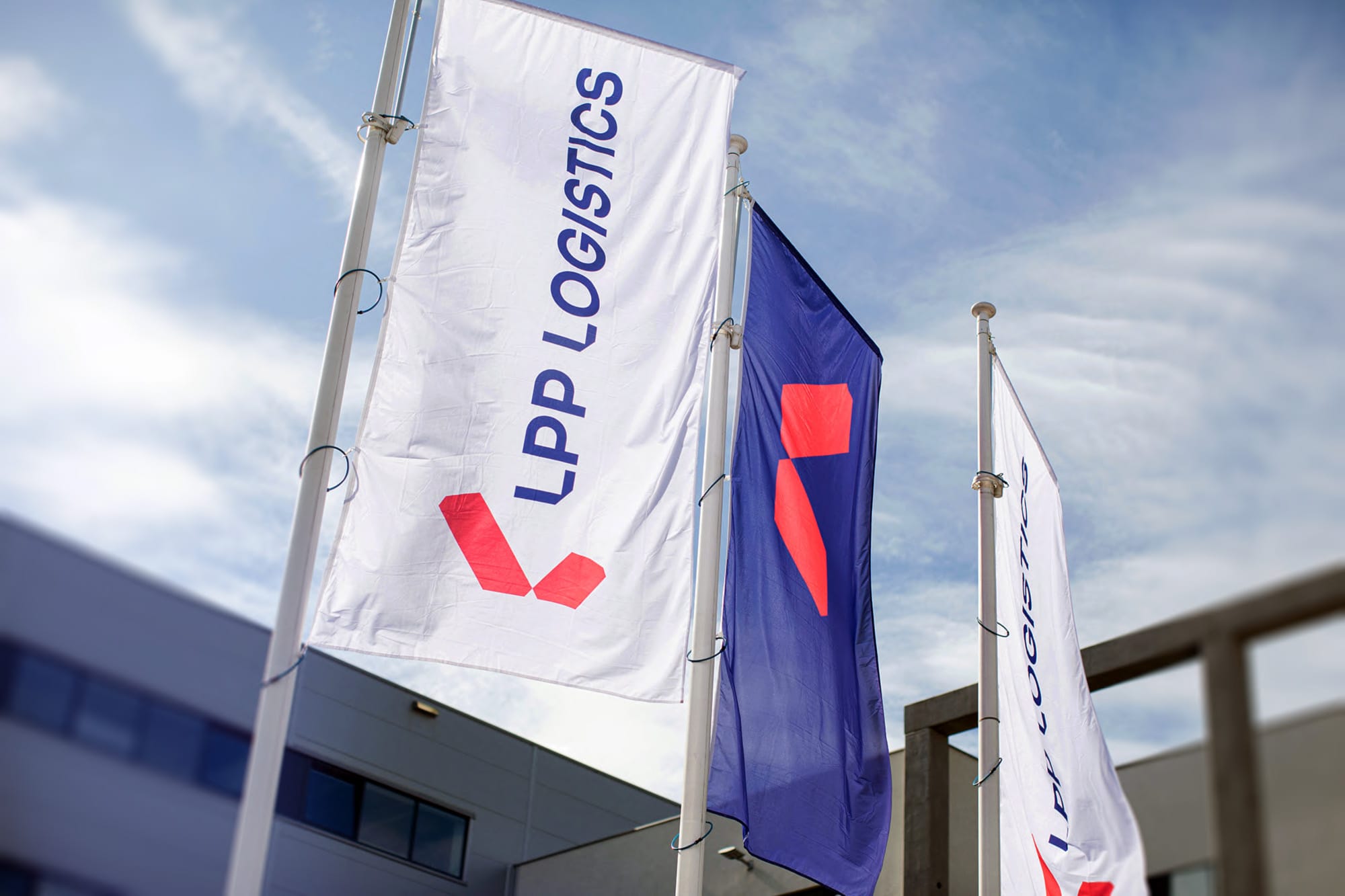 Flagi z logotypem i napisem "LPP Logistics", na tle nieba i budynków