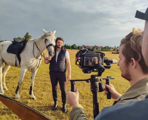 Na zdjęciu ujęcie procesu nagrywania. Na pierwszym planie osoba z kamerą wideo, na drugim planie mężczyzna obok konia.