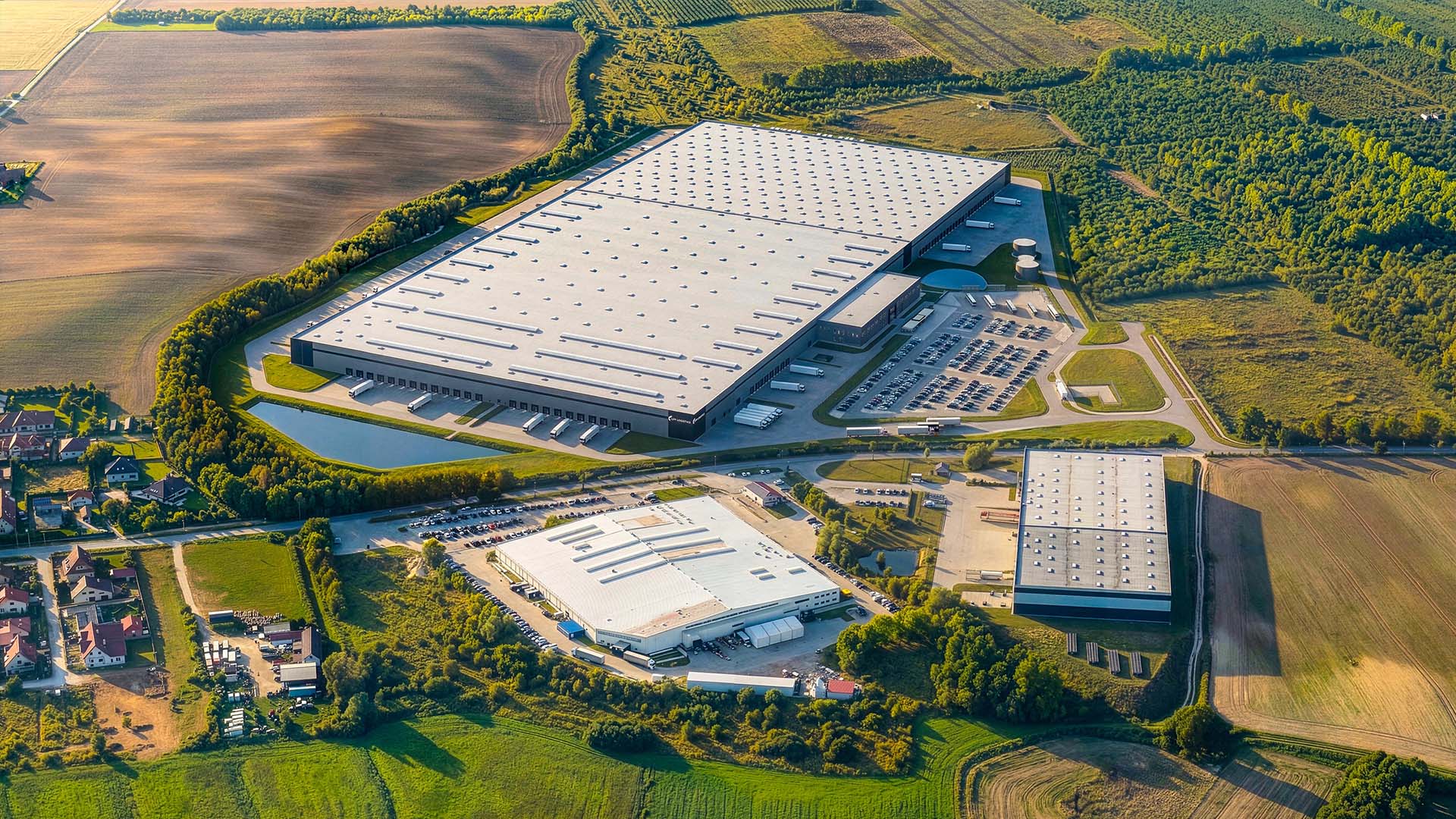 Aerial view of a large industrial warehouse complex with loading docks, parking lots, and surrounding fields and trees in a rural area.