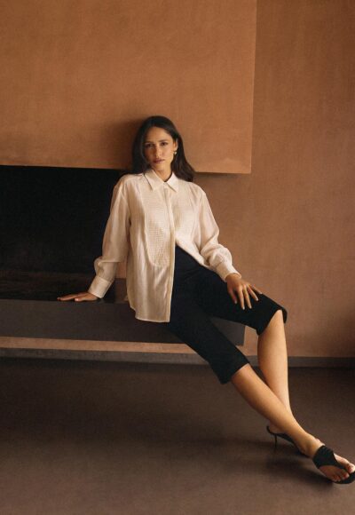 Woman sitting on a dark bench against a brown wall, wearing a white blouse and black pants with heeled sandals.