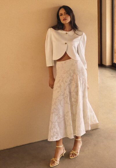 Woman posing in a white cropped blazer over a lace skirt with yellow sandals, against a beige wall.”,