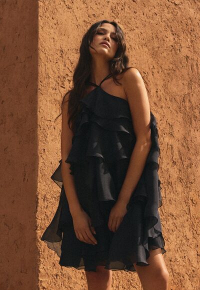 Woman in a black, ruffled dress standing against a sunlit orange stucco wall.
