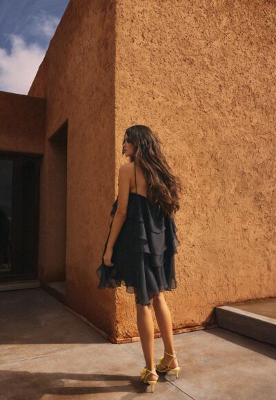 Woman in a black layered dress and yellow heels stands near an orange stucco wall, seen from the back/side outside.