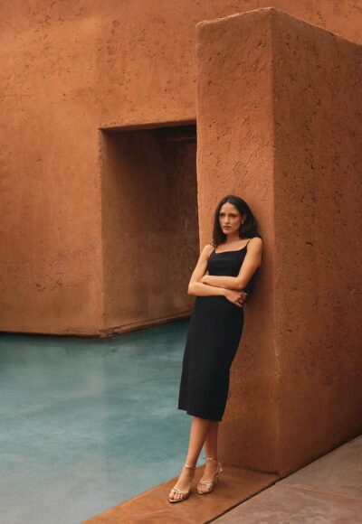 Woman in a black sleeveless dress with strappy sandals leaning against an orange stucco wall by a doorway-shaped opening.