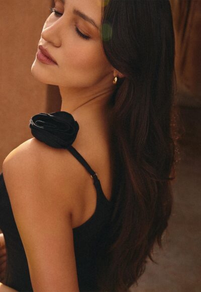 Close-up profile of a woman with long dark hair wearing a black dress with a fabric rose on the shoulder; she is turned slightly away, showing her neck and earrings.
