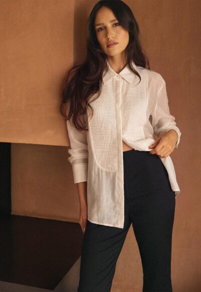 Woman leaning against a warm brown wall in a white long-sleeve blouse and black pants.