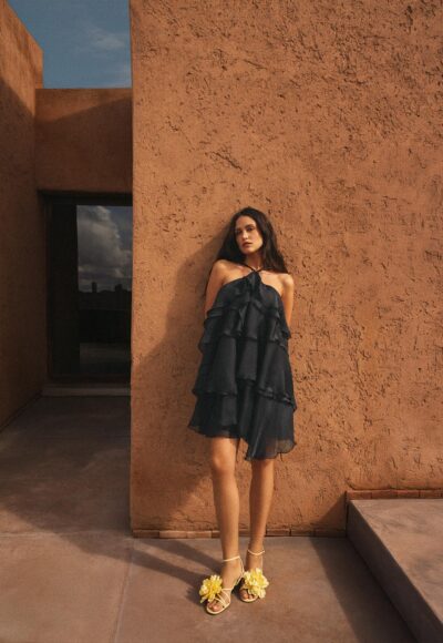 Woman in a black, tiered dress stands against an orange stucco wall, wearing yellow flower-embellished sandals.