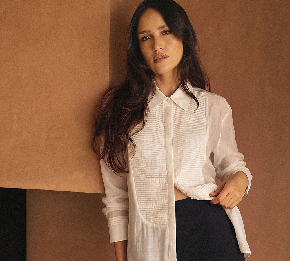 Woman with long dark hair wearing a white pleated-front blouse and black skirt, posing against a tan studio backdrop.