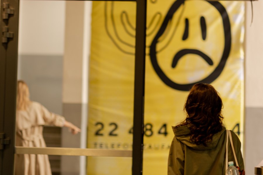 Back view of a person in a green jacket facing a large yellow banner with a sad face; another person points in the background.