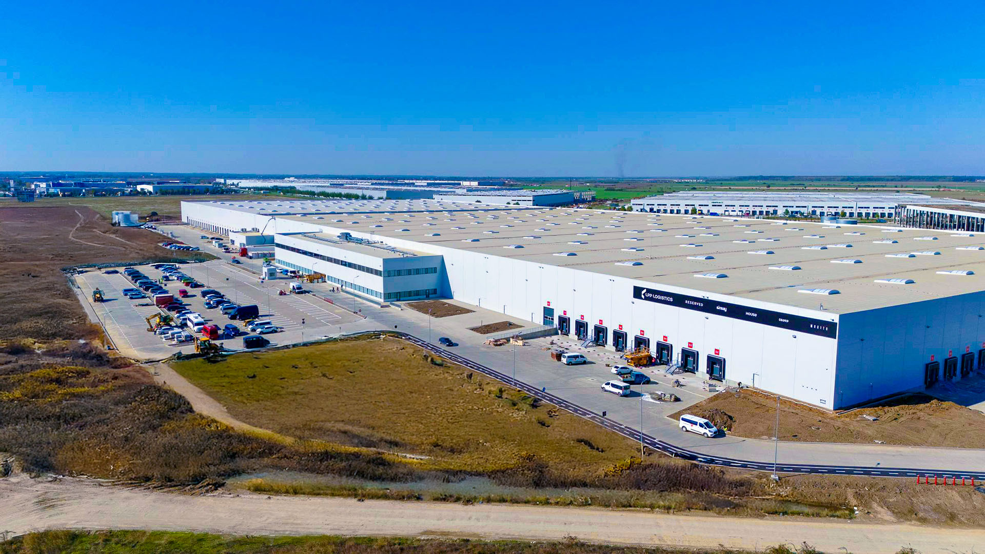 Aerial view of a large white warehouse campus with loading docks, parking lots, and surrounding fields under a blue sky.
