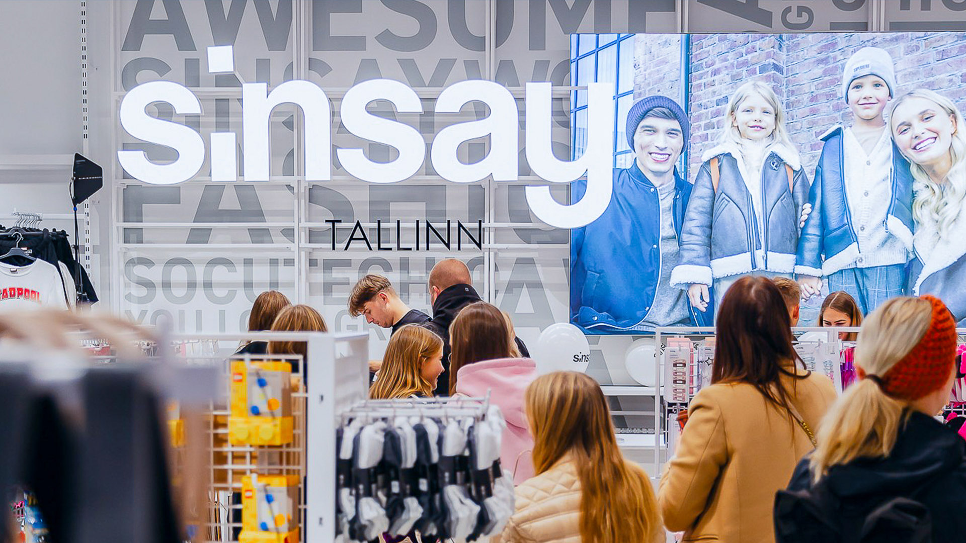 Busy clothing store with a wall mural that reads 'Tallinn' and shows a large family photo; customers browse racks in the foreground.