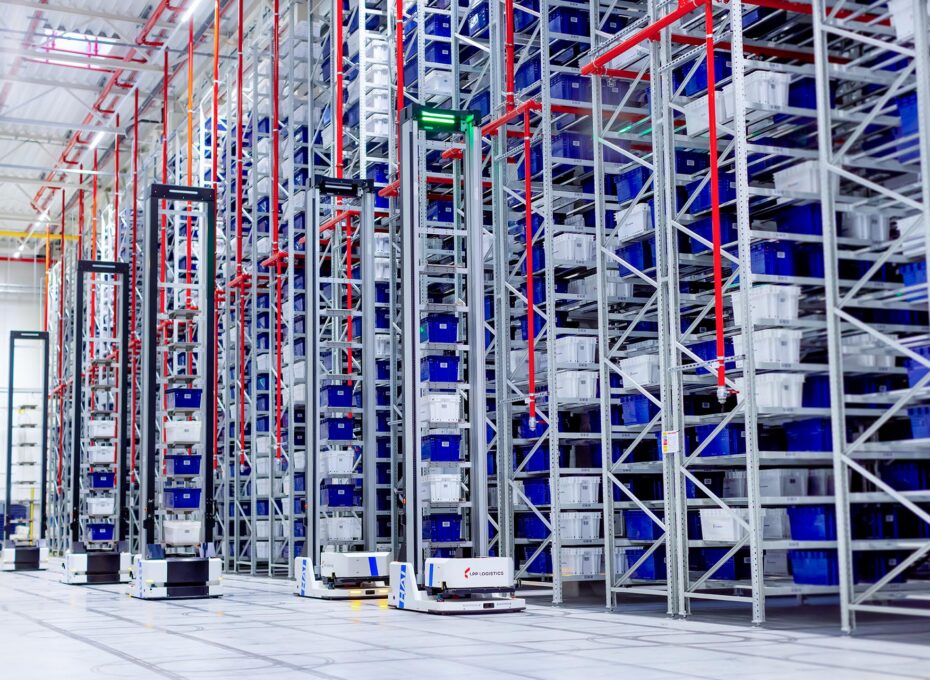 Automated warehouse with tall metal shelving, blue bins, red rails, and small autonomous robots on the floor.