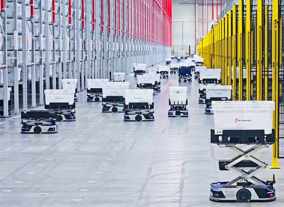 Autonomous delivery robots move through a large warehouse aisle between gray racks on the left and yellow safety barriers on the right, carrying white bins.