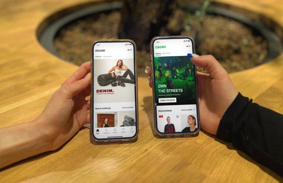 Two hands hold smartphones displaying fashion shopping apps side by side on a wooden table.