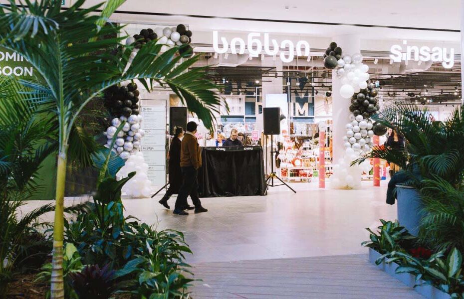 Mall atrium with a balloon-decorated store entrance and two shoppers walking past a DJ booth, greenery in the foreground.