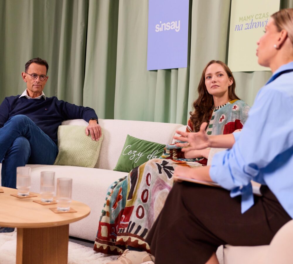 Three people sit on a white sofa in a studio set: a man with glasses on the left, a woman in a colorful dress in the middle, and a moderator in blue on the right, engaged in a discussion with a plant and banners in the background.