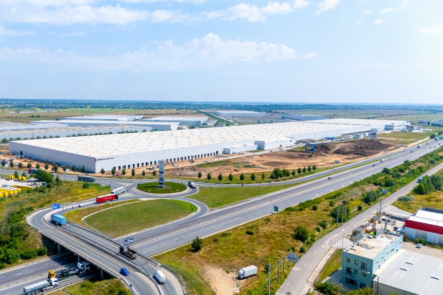Aerial view of a large industrial park with white warehouse buildings and a surrounding highway interchange with multiple lanes and trucks on the roads.