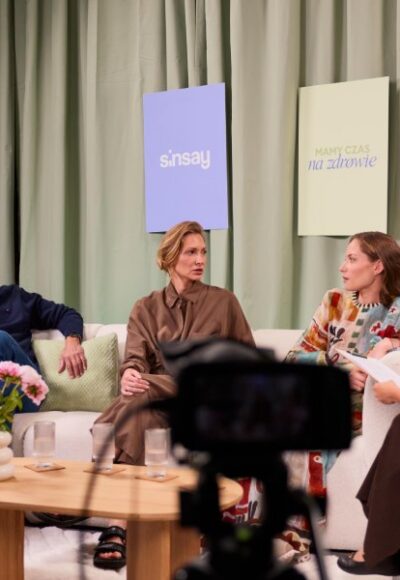 Four panelists sit on a light sofa in a studio, with a camera in the foreground and pastel banners on a green curtain backdrop.