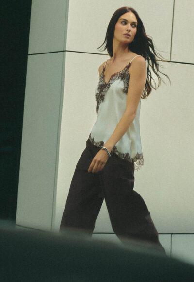 Woman walking outdoors in a white lace-trim camisole and black pants, looking to the side against a modern building backdrop.