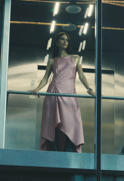 Woman in a pink satin dress stands in a glass elevator, gripping the railing and looking to the side in a moody, urban setting.