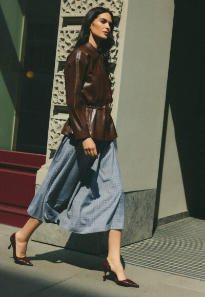 Woman walking past a textured architectural pillar, wearing a brown leather jacket, blue plaid midi skirt, and burgundy high heels.