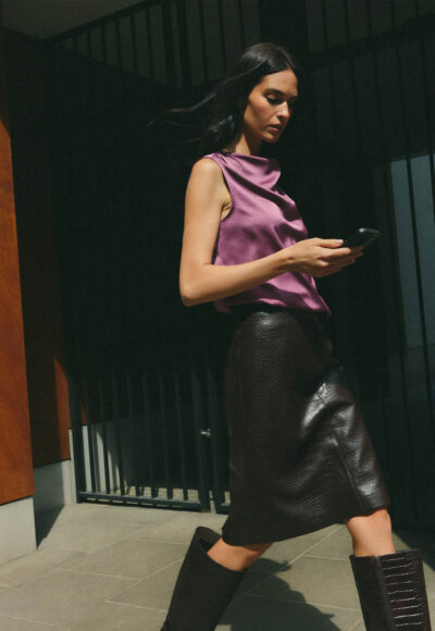 Woman in a magenta satin sleeveless top and black patent-leather skirt walking while looking at her phone.