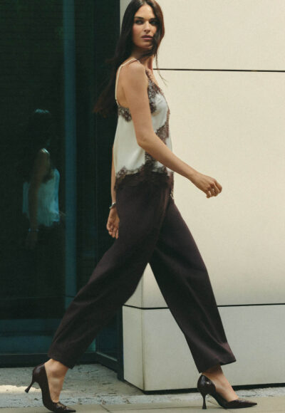 Woman walking in burgundy wide-leg pants and a white lace-trim top, viewed in profile beside a building wall.