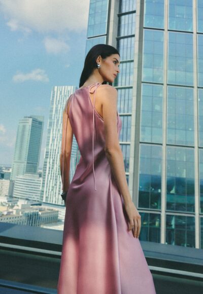 Woman in a pink satin halter dress stands on a rooftop, with glass skyscrapers in the background, looking to the side.