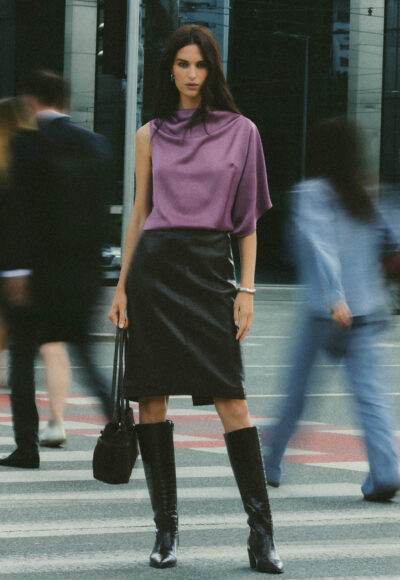 Fashionable woman standing on a city crosswalk in a purple draped top, black leather skirt, and knee-high boots, blurred pedestrians around her.