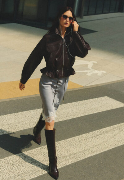 Woman in a black leather jacket, lace-trim denim shorts, knee-high boots, sunglasses, and headphones, walking across a street on a sunny day.