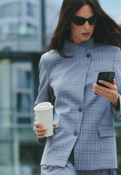 Woman in a light blue plaid blazer holds a takeaway coffee cup in one hand and a smartphone in the other, outdoors with sunglasses on.