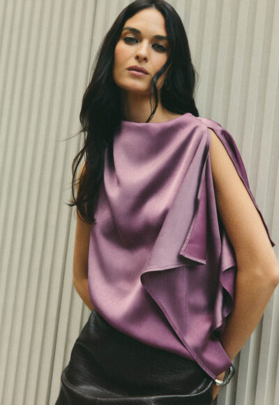 Woman with long dark hair posing against a striped wall in a mauve satin draped top with shoulder cutouts.