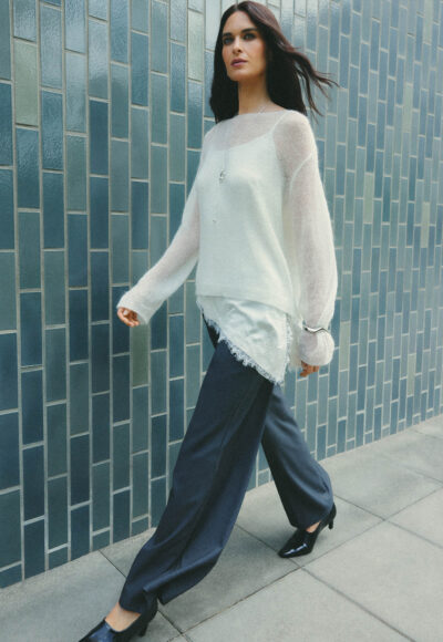 Woman in a white knit sweater and navy wide-leg pants walking beside a blue tiled wall, captured mid-stride by a sidewalk.