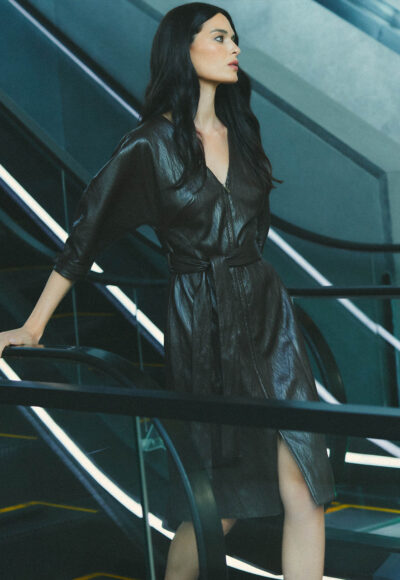 Woman in a black leather dress standing on an escalator in a modern indoor space, looking to the right.