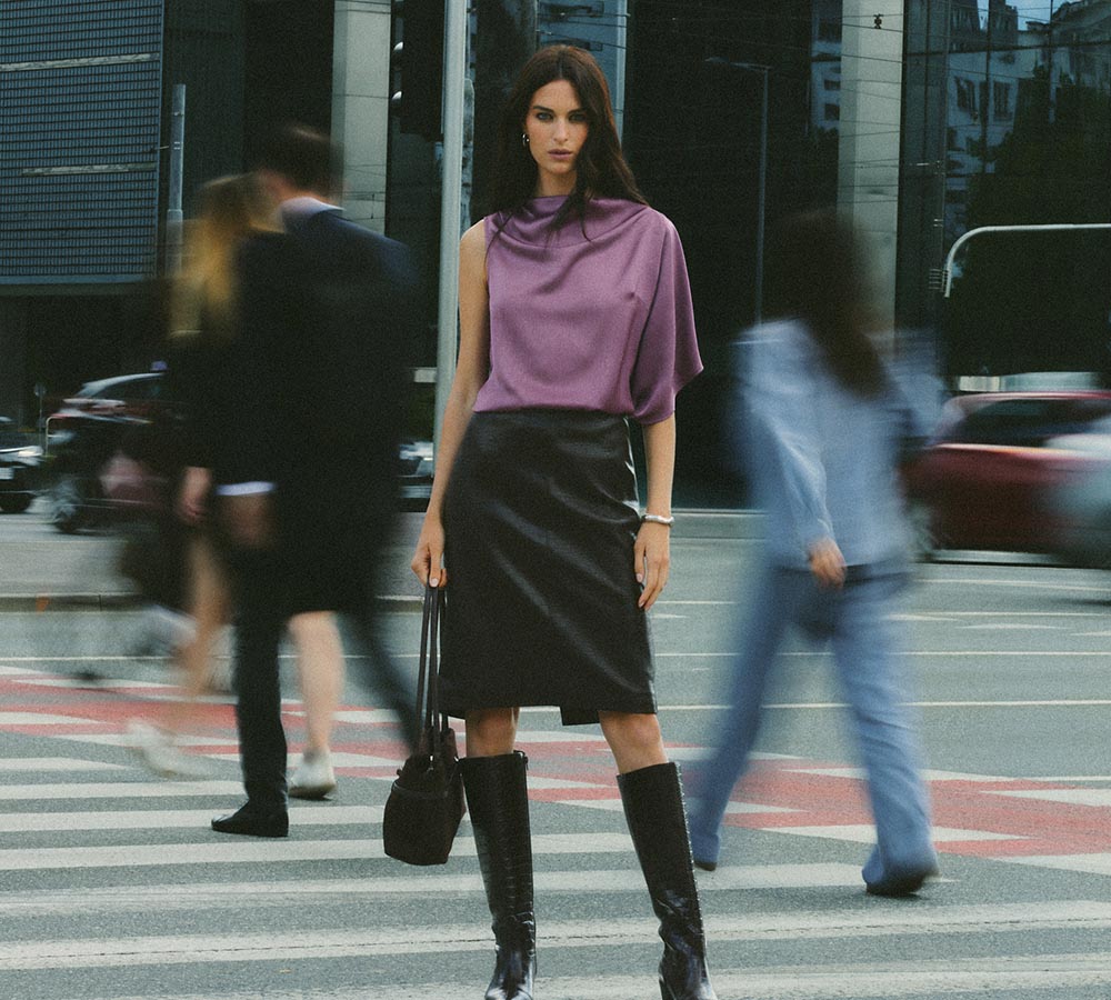 Stylish woman posing at a city crosswalk in a purple draped top, black leather skirt, knee-high boots, and a black bag.