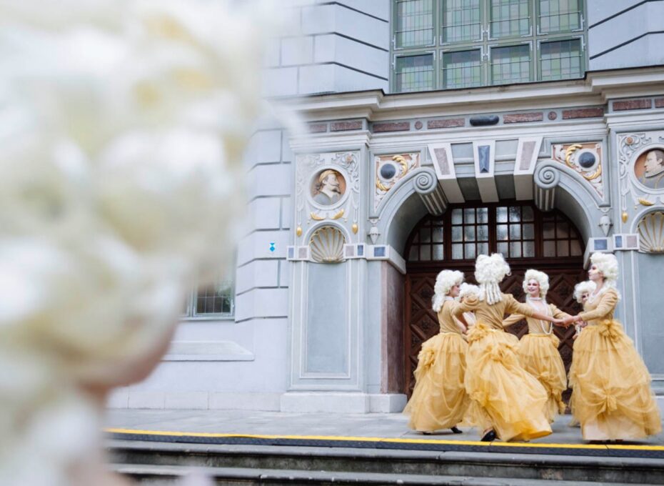 Dancers in yellow 18th‑century gowns and powdered wigs perform in front of an ornate stone building with an arched entrance.