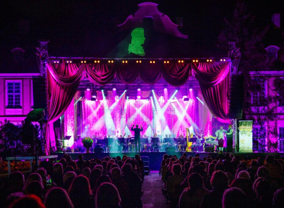Nighttime outdoor concert with a brightly lit stage, purple-pink lighting, curtains, and an audience in front.