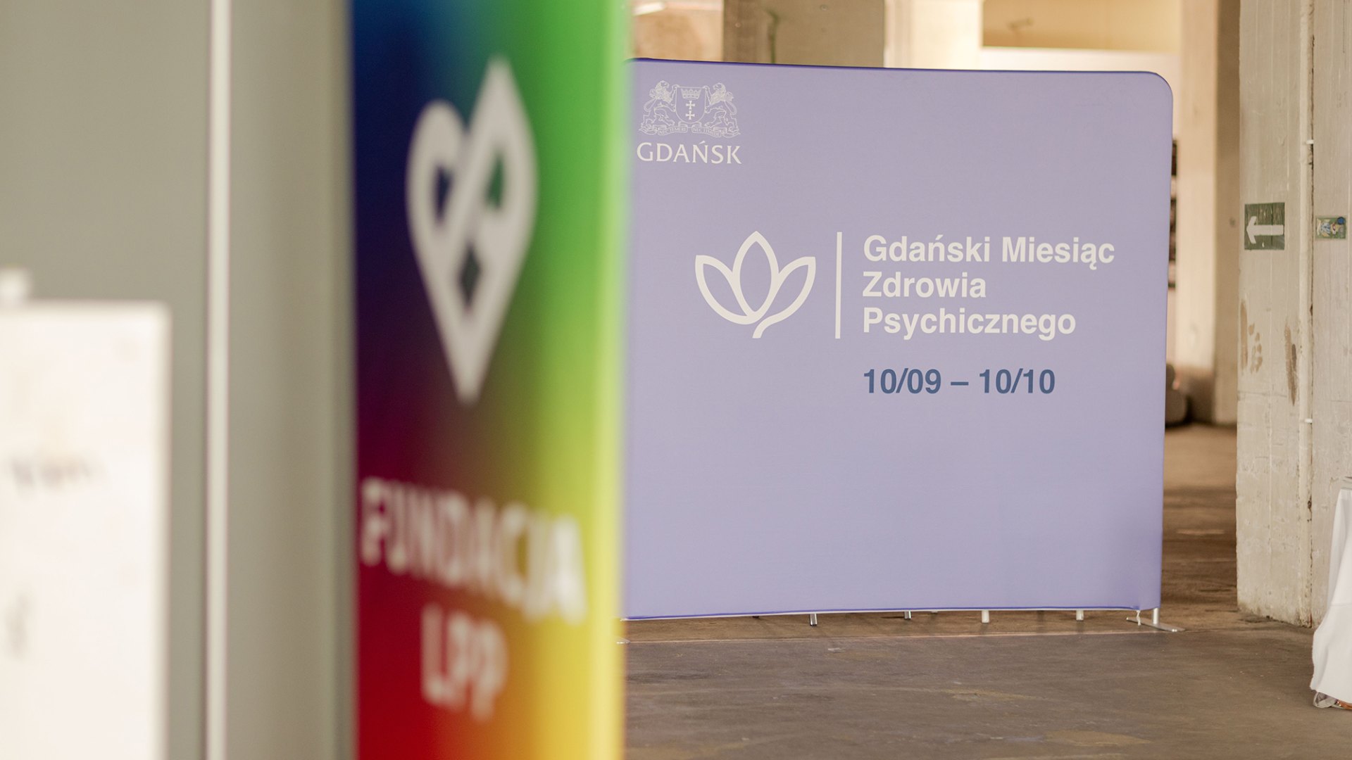Purple sign announcing 'Gdański Miesiąc Zdrowia Psychicznego' with a lotus icon and dates 10/09–10/10 in a hallway.