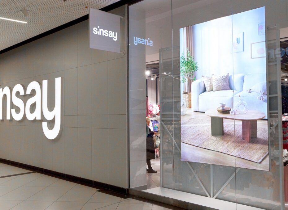 Storefront of a Sinsay shop with a large white logo on a gray wall and glass display windows showing a living room ad.