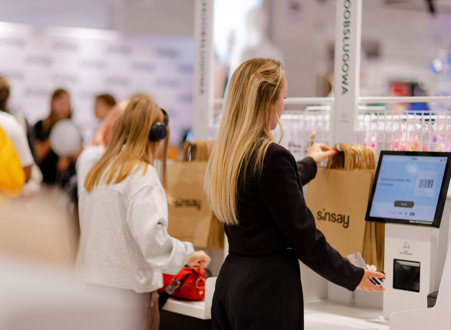 Blonde woman at a self-checkout kiosk tapping the screen, with shoppers and bags in the busy store in the background.