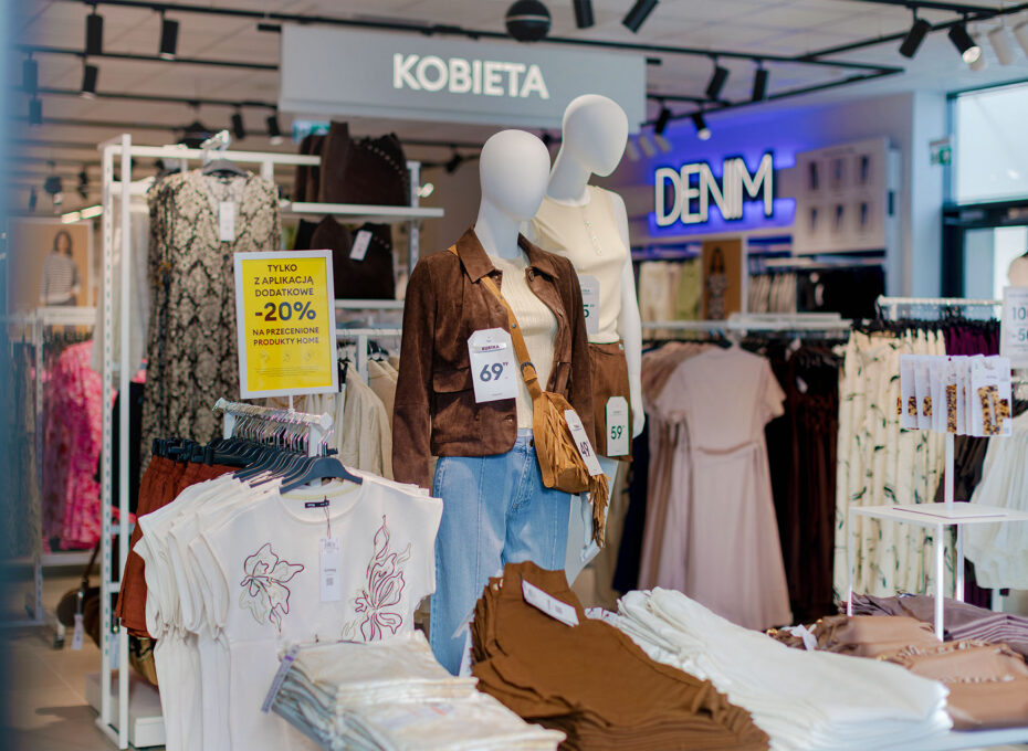 Women’s clothing store display with mannequines wearing brown suede jacket and beige top, surrounded by racks of garments; signs read KOBIETA and DENIM in the background.