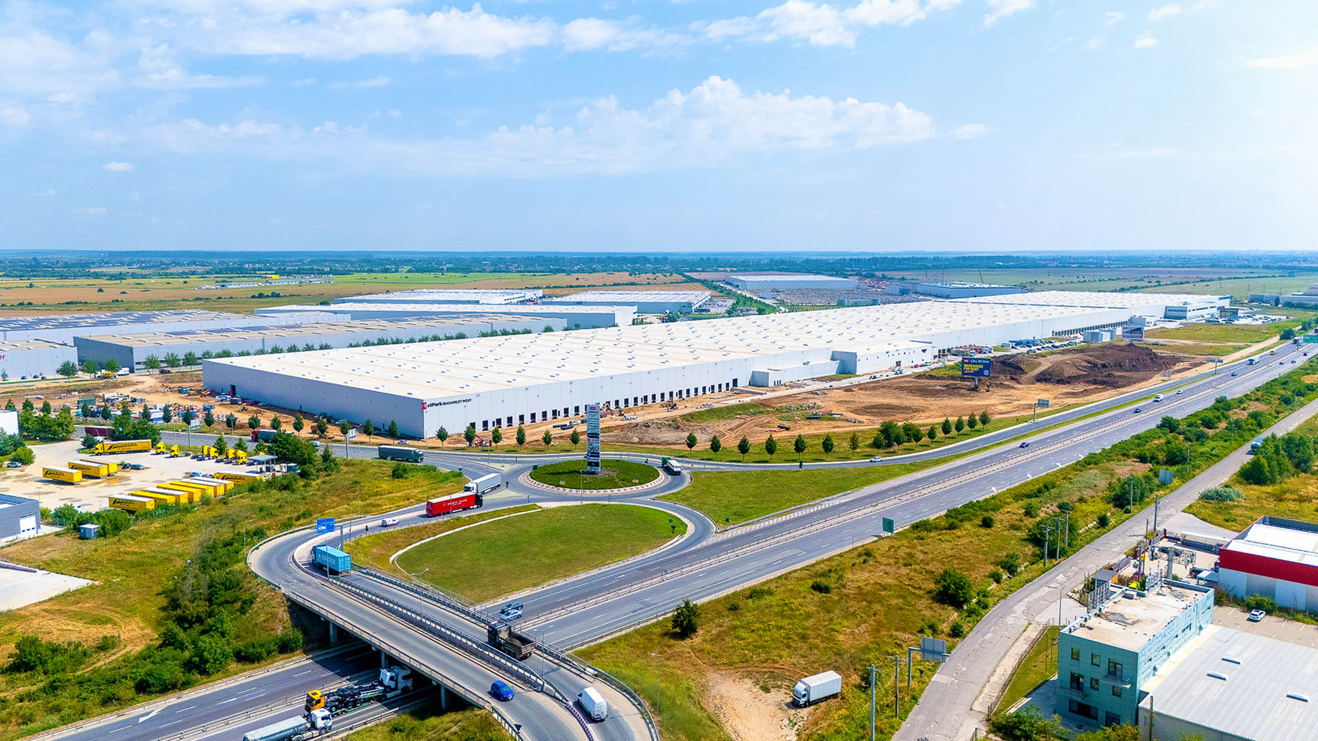 Aerial view of a large industrial park with white warehouse buildings, a roundabout, and highway junctions with trucks and cars.