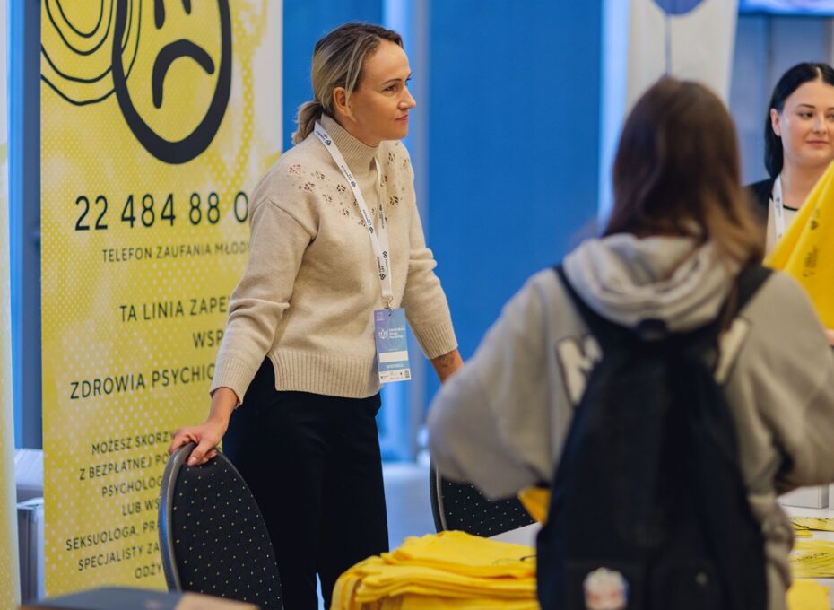 Staff member in a beige sweater with a lanyard helping visitors at a bright yellow information booth.