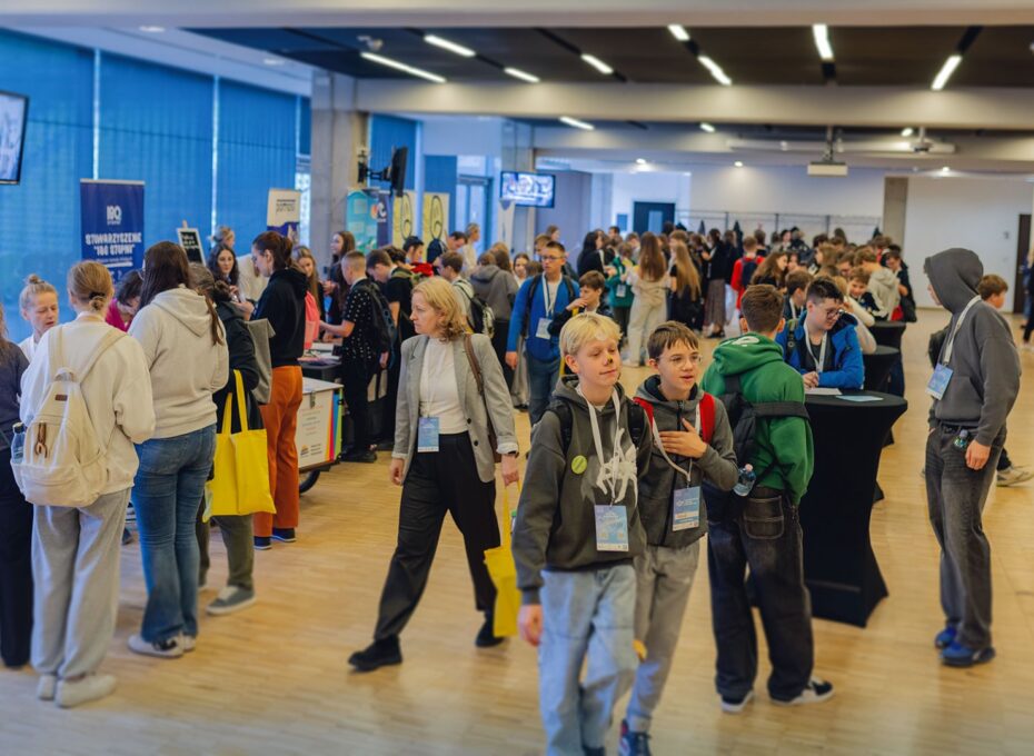 Crowded indoor expo with attendees chatting at booths and carrying yellow tote bags and lanyards.
