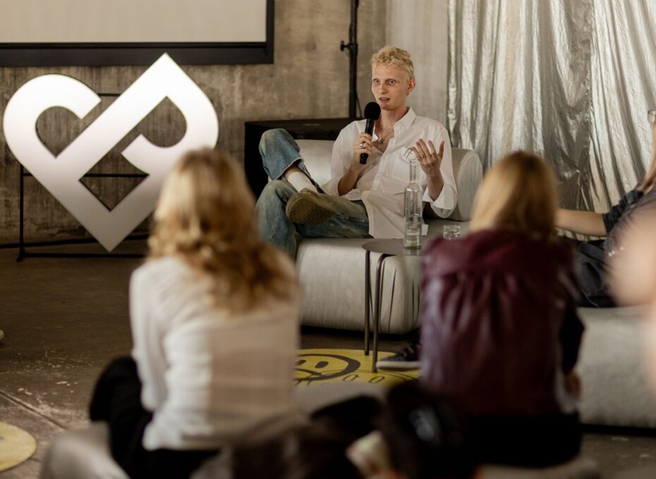 Person with short blond hair in a white shirt sits on a white chair, speaking into a microphone to a small audience in a casual panel setup.