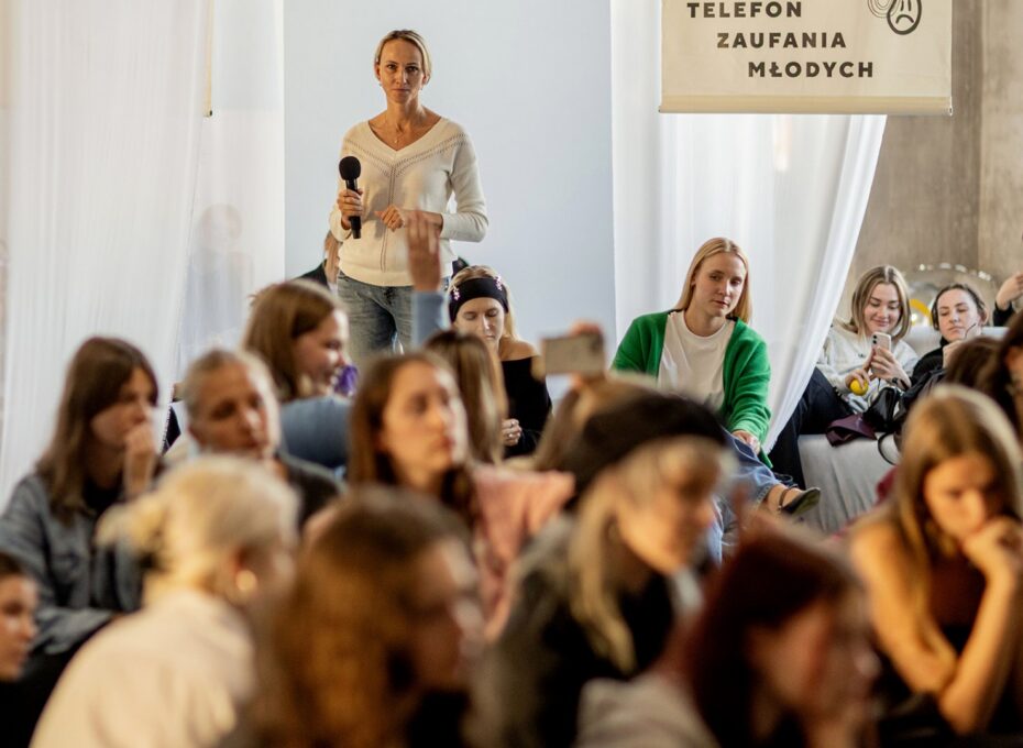 Woman standing with a microphone addressing a crowd at a youth event in a bright room with a banner overhead.
